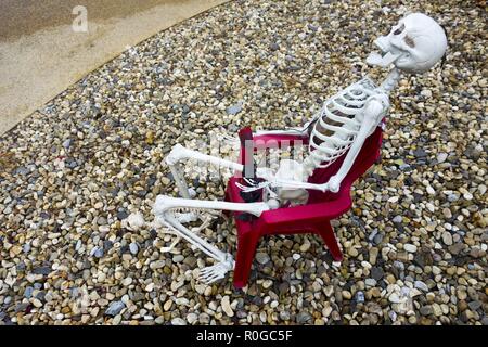 White Human Skeleton Profile Model Side View sitting in Backyard Red Adirondack Chair Halloween Decoration Stock Photo