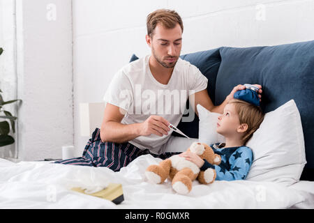 father checking sons temperature with electric thermometer and holding ice pack on his head in bed Stock Photo