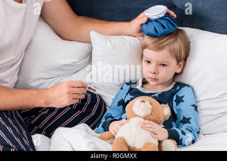 cropped shot of father checking temperature of son with electric thermometer and holding ice pack on his head in bed Stock Photo