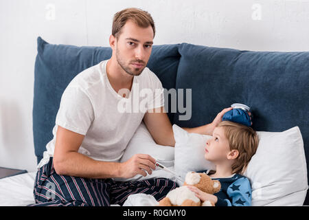 father checking temperature of son with electric thermometer and holding ice pack on his head in bed and looking at camera Stock Photo