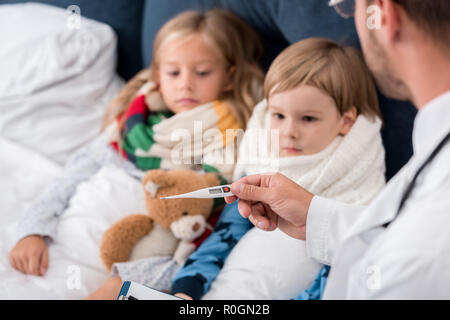 pediatrician with clipboard examining temperature of kids lying in bed Stock Photo