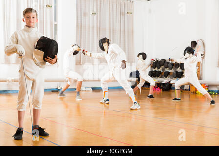Portrait of glad boy wearing fencing uniform standing in gym with foil and mask in hands Stock Photo