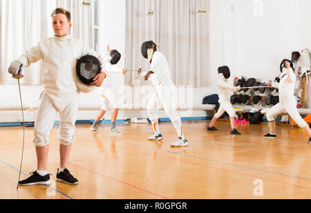 Portrait of happy boy wearing fencing uniform standing in gym with foil and mask in hands Stock Photo