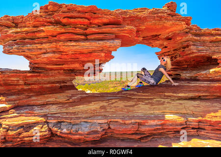 Blonde happy woman sitting inside the iconic rock arch in red sandstone of Nature's Window, Kalbarri National Park. Caucasian young girl enjoying in Western Australia. Australia travel freedom concept Stock Photo