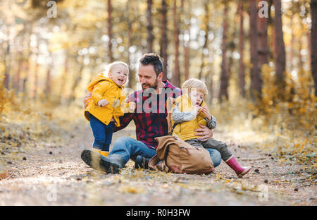 A mature father with toddler children sitting on the ground in an autumn forest. Stock Photo