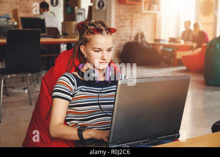 Hipster cute student girl with dreads wearing headphones using laptop at the coworking Stock Photo