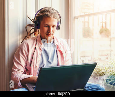Hipster attractive bearded man with dreads wearing headphones sitting near the window and chatting in the social networks Stock Photo