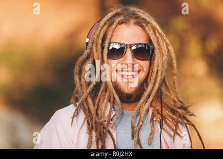 Portrait of hipster blonde bearded boy with dreads wearing headphones and glasses walking and smiling outdoors an the sunny autumn day Stock Photo