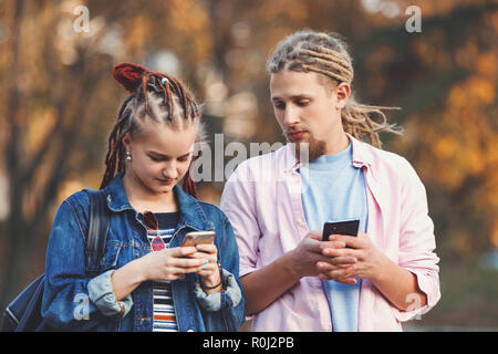 Two hipster best friends with dreads wearing casual clothes typing messages on their smartphones, bearded blonde man looking on the smartphone of his girlfriend, outdoor view Stock Photo