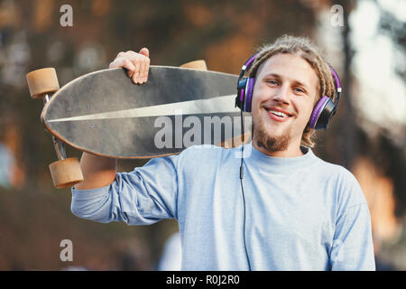 Hipster skater blonde bearded man with dreads wearing headphones laughing and walking in the forest, outdoor view Stock Photo
