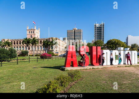 Five tourists are posing with 'ASUnción' letters and Palacio de los López (Presidential palace) in background. Asuncion city, Paraguay, Latin America Stock Photo