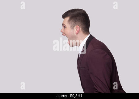 Profile side view portrait aggressive of handsome young man in violet suit and white shirt, standing and screaming with nervous face. indoor studio sh Stock Photo