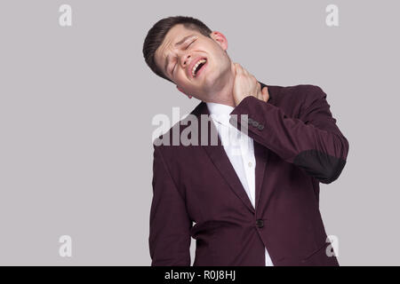 Portrait of painful sick handsome young businessman in violet suit and white shirt, standing with closed eyes and feeling pain on his neck. indoor stu Stock Photo