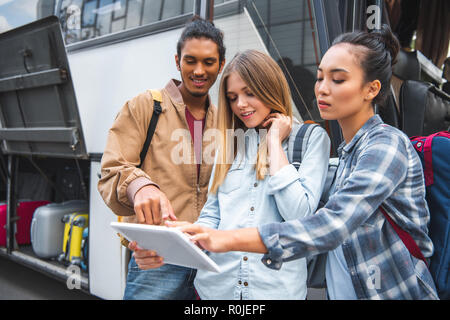 mixed race young man pointing at digital tablet to female friends near travel bus at city street Stock Photo