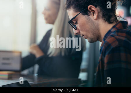 Close up of young man wearing eyeglasses sitting in meeting at office. Young executive in casuals in meeting with colleagues sitting around. Stock Photo