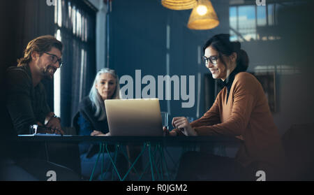 business professionals sitting around table and looking at laptop. Asian female executive giving a presentation over laptop to colleagues at office. Stock Photo
