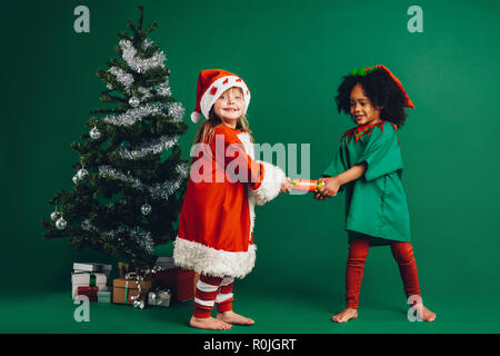 Two little girls in christmas costumes pulling a toy. Kids having fun playing with a toy standing beside a decorated christmas tree. Stock Photo