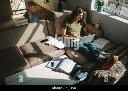 Student studying for exams sitting on couch with sunlight coming from window. Student studying hard on laptop and books. Stock Photo