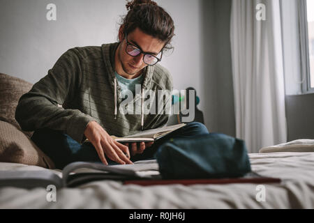 Student sitting on a bed and studying with focus and concentration. Young man preparing for exam reading books. Stock Photo