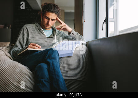 Student sitting at home looking tensed studying seriously. Man sitting on couch beside a window writing in a book. Stock Photo