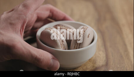 man hand put beige macarons in white bowl on wood table, wide photo Stock Photo