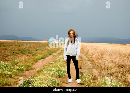 Beautiful young woman red-haired posing in a path between two wheat field. Cloudy day. Teenager Stock Photo