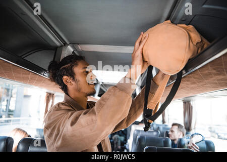 side view of young mixed race man putting rucksack on shelf in travel bus Stock Photo