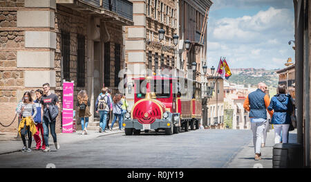 Toledo, Spain - April 28, 2018: Tourist strolling in a street of the historic district next to a small tourist red train on a spring day Stock Photo