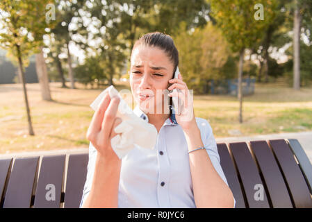 Sad young woman cries while talking on the mobile phone in the park on bench Stock Photo