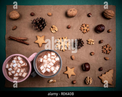 Above view with two cups with mini marshmallows and hot chocolate on a brown tablecloth decorated with nuts and winter sweets
