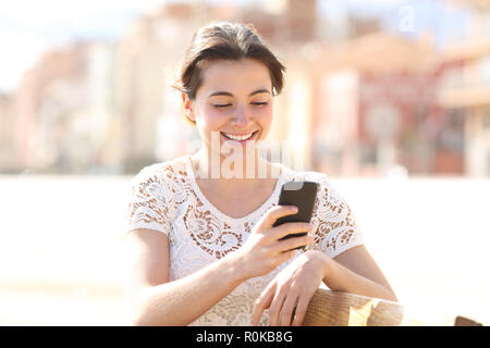 Happy lady uses a smartphone sitting on a bench in a park Stock Photo