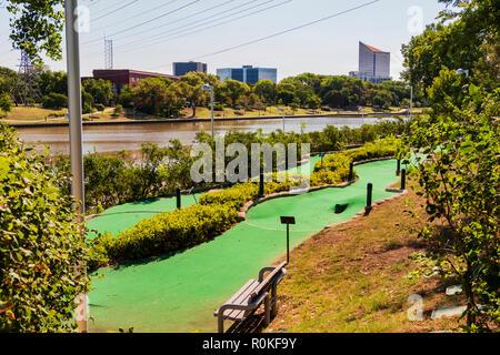 A minature golf course running beside the Arkansas river with city buildings  in background. River Walk on opposite shore. Wichita, Kansas, USA. Stock Photo