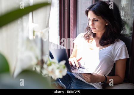 Attractive young woman relaxing on a sunny window seat with a laptop enjoying the sunshine with green houseplants in the foreground Stock Photo