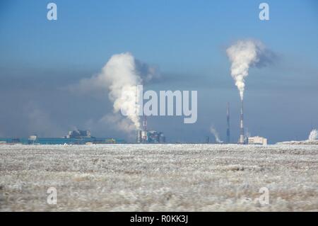 Winter landscape with metallurgical plant with heavy smoke from pipes behind a field covered with frozen dry grass under dark blue sky Stock Photo