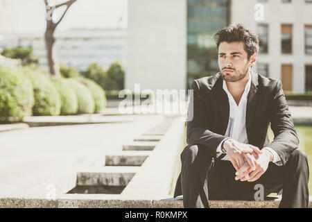 Handsome young man sitting in the city. Relaxed thinking with his arms on her legs. Business suit with black jacket and trousers. Stock Photo
