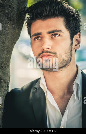 Handsome young man thinking. Outdoors, near a tree. The young man is wearing a white shirt and a jacket. Looking upwards. Stock Photo