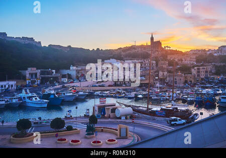 GHAJNSIELEM, MALTA - JUNE 15, 2018: The scenic fishing harbor with boats, rocky hills and tall silhouette of Parish Church on twilight, on June 15 in  Stock Photo