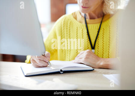 Hand of mature businesswoman making notes in notepad by her workplace Stock Photo