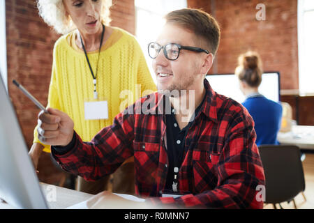 Young smiling analyst pointing at data on computer screen while explaining it to mature employer Stock Photo