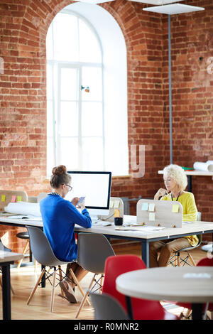 Young and mature colleagues sitting by their workplaces in office and using modern technologies Stock Photo