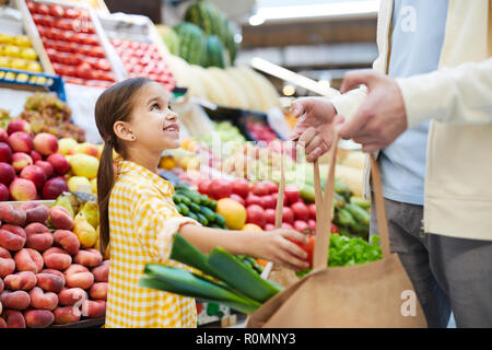 Cheerful cute girl in yellow dress putting vegetable into bag and looking at father while helping him with shopping at farmers market Stock Photo