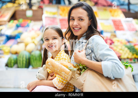 Cheerful beautiful family doing shopping at farmers market: smiling attractive mom with bag full of fresh product embracing cute daughter and looking  Stock Photo