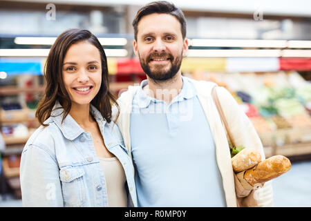 Happy positive young couple embracing and looking at camera while standing in food shop, smiling bearded man holding shopping bag on shoulder Stock Photo