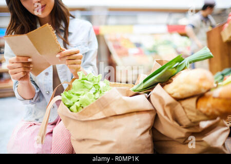 Close-up of content young woman crouching near full shopping bags and reading shopping list in organic food store Stock Photo