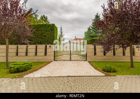 Iron gate with trees and stone fence, luxury house or villa guard. Elegant driveway towards luxury villa Stock Photo
