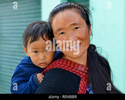 Young Indian Monpa woman carries her toddler boy in a traditional baby sling on her back. Stock Photo
