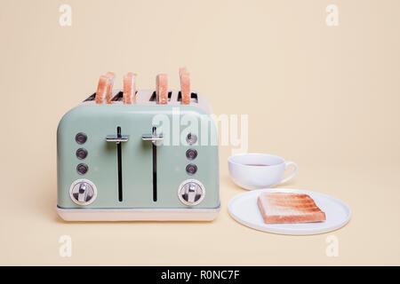 Retro green four slice double toaster filled with toast and a piece of toast on a white plate with a cup of coffee on a vintage tan background Stock Photo