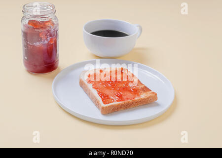 Slice of toast with strawberry jam jelly on a white plate with a cup of coffee and a jar of preserves on a vintage tan background with copy space Stock Photo