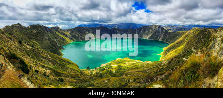 A bird's eye panoramic view of the bright green volcanic Quilotoa Lake in Ecuador with lots of white and grey clouds in a blue sky and green brush on  Stock Photo