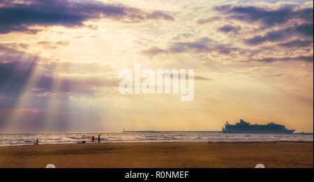 Horizontal photo of the rays from the sun low on the horizon silhouetting a cruise ship and people and dogs on the shore of a sandy beach